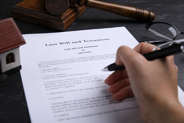 Person signing a Last Will and Testament document at a desk with a gavel, glasses, and a small house model nearby.