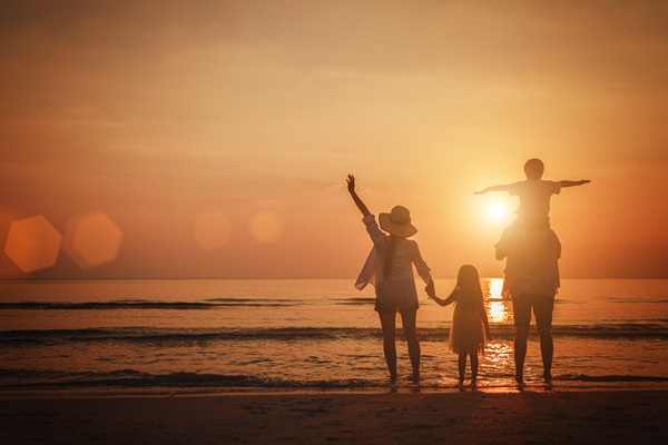 A family of four stands on the beach at sunset, holding hands and enjoying the ocean view. The sun sets over the horizon, casting a warm glow over the scene, with one parent carrying a child on their shoulders.