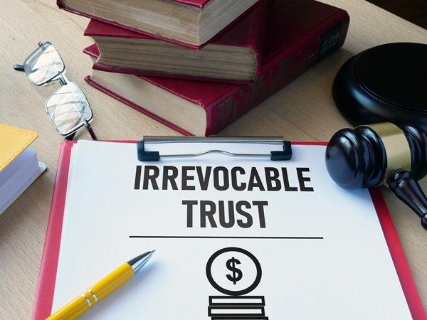 A white document with the words "IRREVOCABLE TRUST" printed in large, bold black letters sits on a red clipboard alongside a yellow pen. In the background, there are three red hardcover books, a pair of eyeglasses, and a wooden judge's gavel.