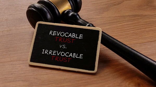 A gavel rests on a wooden surface next to a small chalkboard sign reading "REVOCABLE TRUST VS. IRREVOCABLE TRUST."